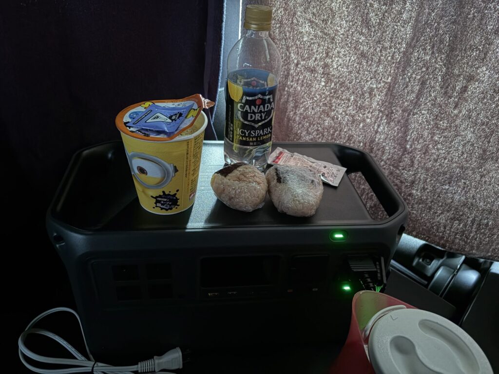 バスタオルカーテンで暗くなった車内で、ポータブル電源の上に置かれたおにぎりとカップ麺